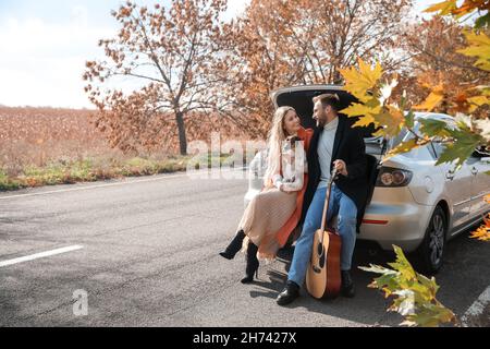 Lovely young couple with cute dog and guitar sitting in car trunk outdoors Stock Photo