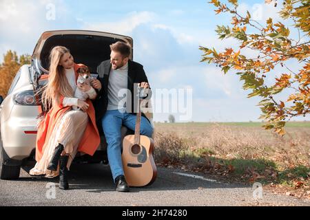 Lovely young couple with cute dog and guitar sitting in car trunk outdoors Stock Photo