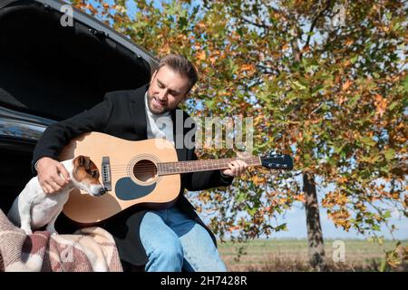 Handsome man with cute dog playing guitar in car trunk on autumn day Stock Photo