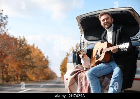 Handsome man with cute dog playing guitar in car trunk on autumn day Stock Photo