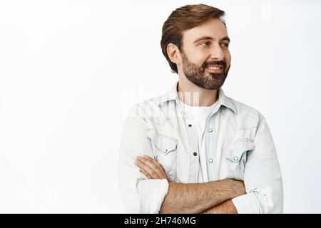Close up portrait of handsome caucasian man looking sideways and smiling, standing in casual pose with crossed arms, white background Stock Photo