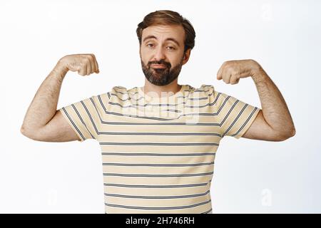 Image of sad funny guy showing his muscles, flexing biceps and grimacing upset with his body, standing over white background. Concept of workout and Stock Photo