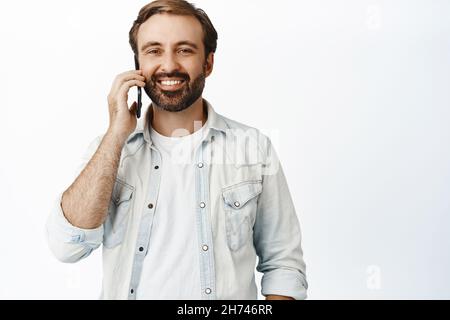 Handsome casual guy talking on phone, calling someone and smiling, standing against white background Stock Photo