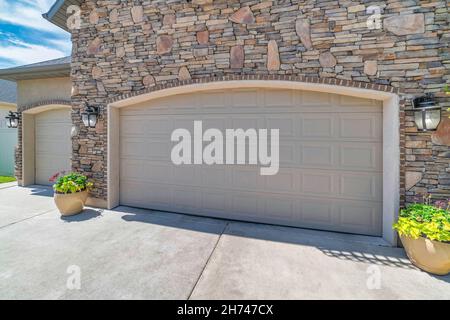 Two beige sectional garage doors with arched bricks door opening. Exterior of a three car garage with stone veneers wall and two potted plants at the Stock Photo