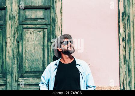 Hipster tourist smile at the sun standing against an old wall with green door and window. Happy adult mature man people with hat and sunglasses enjoyi Stock Photo