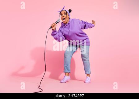 Photo of pretty shiny african lady dressed violet sweatshirt buns smiling singing songs isolated pink color background Stock Photo