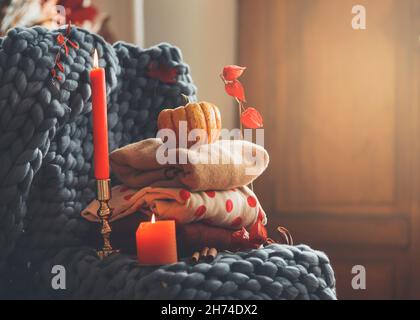 Cozy autumn still life with knitted sweaters, candles, grey wool blanket, candles and autumn leaves at interior background. Warm clothes for autumn se Stock Photo