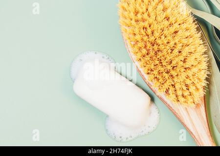 Soap bar with foam bubbles next to massage brush on green background. Hygiene and body care concept, flat lay Stock Photo