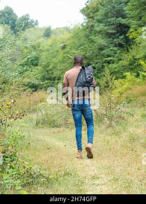 Back view of an African tall man in stylish clothes with a backpack walking on the grassy pathway Stock Photo