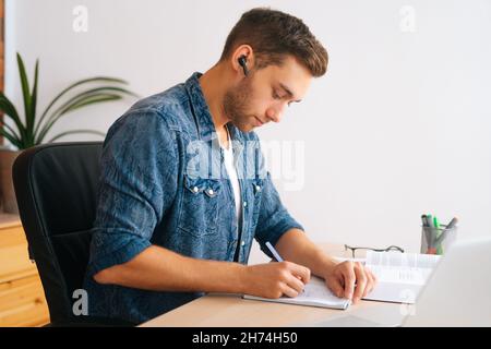 Side view of concentrated male student studying online from home using laptop writing notes, watching video class lesson, listening audio course. Stock Photo