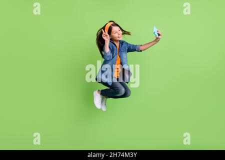 Full size profile photo of cool small girl jump do selfie wear hoop jeans shoes isolated on green background Stock Photo