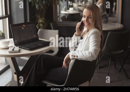 Business woman talking on the phone, smiling, working with laptop in cafe with dark black interior Stock Photo