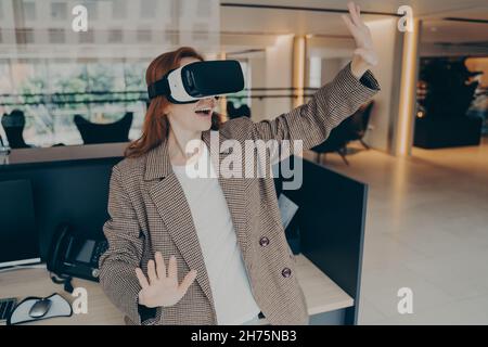 Woman wearing VR headset surprised with virtual reality while standing near her workplace in office Stock Photo