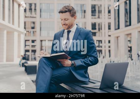Relaxed confident office worker in formal blue suit writing notes in agenda while working outside Stock Photo