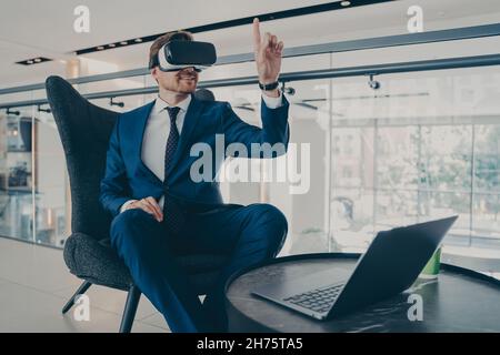 Well-groomed CEO man in formal suit wearing VR headset goggles, while sitting in office lobby Stock Photo