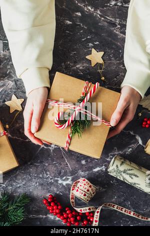 Womans hand holding Christmas gift box over slate desk table. Vintage Christmas present decorated red and white striped ribbon, fir branch, candy cane Stock Photo