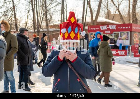 A girl in a buffoon's cap looks at a mobile phone among people at the holiday Seeing off the Winter in the city park. Stock Photo