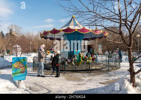A family, along with dad with a child on their shoulders, stand near a carousel for toddlers in a city park for recreation and entertainment among the Stock Photo