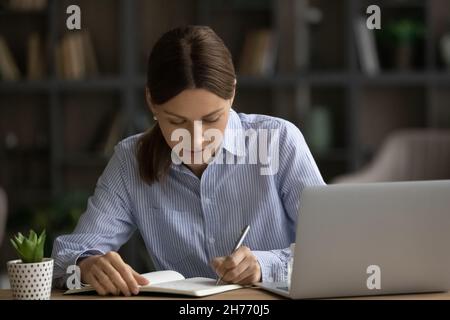 Concentrated millennial woman sit at desk jotting information in notebook Stock Photo