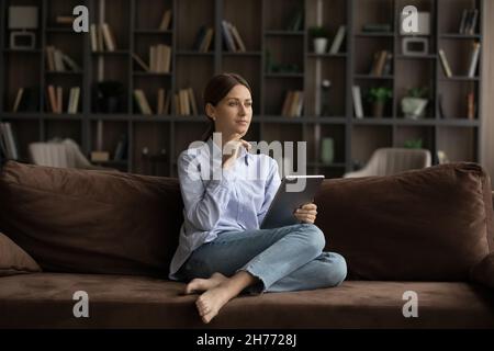 Pensive woman sit on sofa with digital tablet Stock Photo