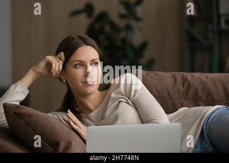 Dreamy woman lying on sofa with laptop looking into distance Stock Photo