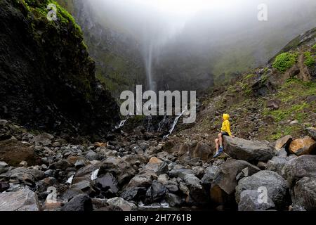 Boy sitting on large rock at the bottom of Ribeira grande waterfall. Flores, Azores, Portugal Stock Photo