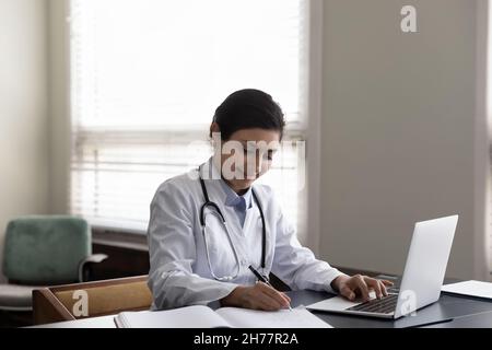 Smiling Indian woman doctor taking notes, using laptop in office Stock Photo