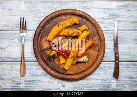 Roasted pumpkin slices and baked vegetable on wooden plate. Table top view of place setting with rustic cutlery. Tasty vegetarian food Stock Photo
