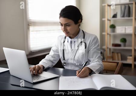 Serious Indian woman doctor physician using laptop, taking notes Stock Photo