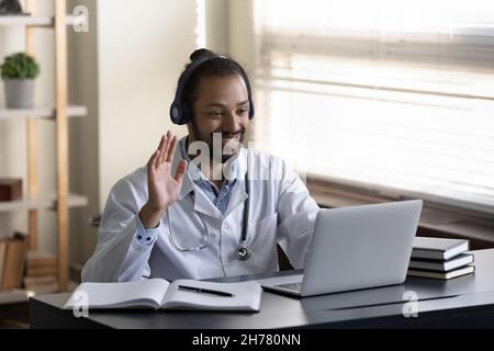 Smiling African American doctor in headphones using laptop, waving hand Stock Photo