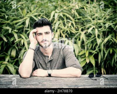 Attractive young man in city park in a nice summer day, sitting on wood bench, leaning on table Stock Photo