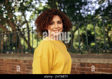Portrait of a young teenage girl smiling cheerfully outdoors. Cheerful female youngster wearing red hair and a mustard sweatshirt in the city. Young t Stock Photo
