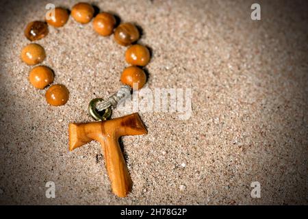 Tau, wooden cross in the shape of the letter t (symbol of St. Francis of Assisi) with rosary bead partially buried in the sand Stock Photo