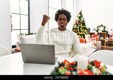 Young african american man using laptop sitting on the table by christmas tree angry and mad raising fist frustrated and furious while shouting with a Stock Photo