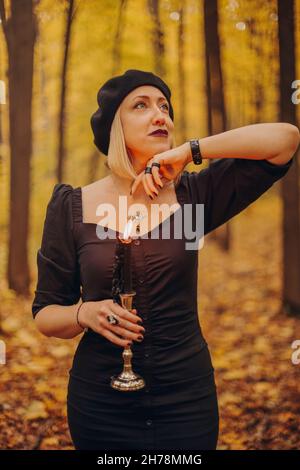 Shot of mysterious blond woman in black dress holding candle on candlestick in her hands performing magic ritual in dark autumn forest. Halloween conc Stock Photo