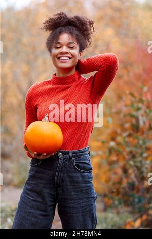 Young mixed-race woman with curly hair holding orange pumpkin while standing against blurred autumn forest background. Fall season and harvest concept Stock Photo