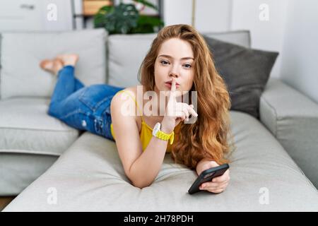 Young caucasian woman lying on the sofa using smartphone asking to be quiet with finger on lips. silence and secret concept. Stock Photo