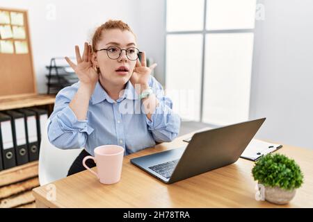Young redhead woman working at the office using computer laptop trying to hear both hands on ear gesture, curious for gossip. hearing problem, deaf Stock Photo