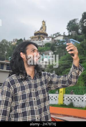 A charming long haired and bearded young guy smiling with taking selfie or having video call at outside with his mobile phone Stock Photo
