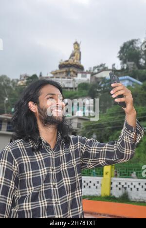 A charming long haired and bearded young guy smiling with taking selfie or having video call at outside with his mobile phone Stock Photo