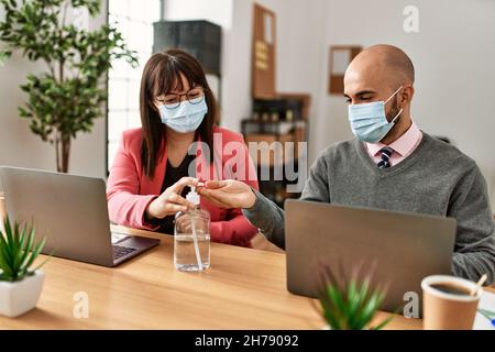 Two hispanic business workers wearing medical mask using sanitizer hands gel at the office. Stock Photo