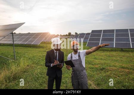 Indian technician with digital tablet in hands showing solar plant to african american inspector with clipboard. Two men working on producing alternative energy. Stock Photo