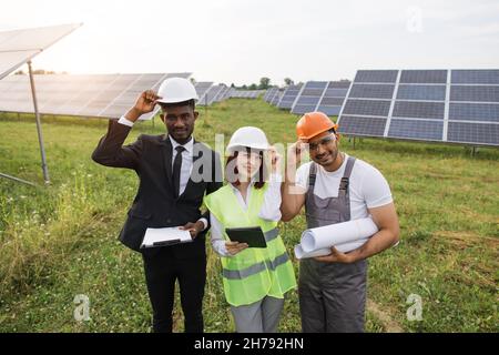 African american businessman, caucasian woman and indian foreman having meeting about solar energy. Multicultural people touching their safety helmets on head and smiling on camera. Stock Photo
