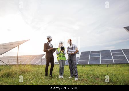 Indian technician in yellow helmet standing at solar plant with two inspectors, smiling and looking around. Male and female engineers using clipboard and tablet for work. Stock Photo
