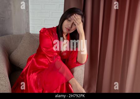 Asian woman in a red satin nightgown and red long full length robe with headache, she is sitting on the gray sofa and touching her forehead. Thai woma Stock Photo
