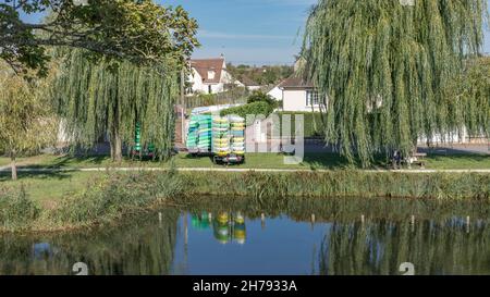 Different coloured Kayaks piled up by the side of a river surrounded by trees and under a blue sky Stock Photo