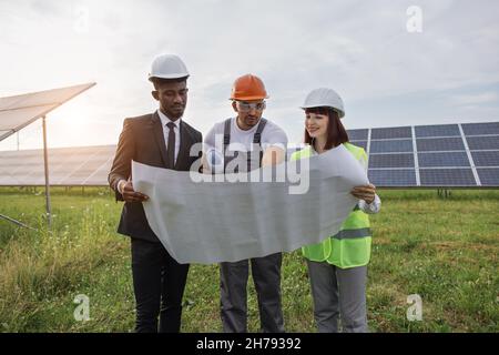 Two engineers and one technician examining drawings of solar farm while standing together outdoors. Male and female partners in helmets working on modern business project. Stock Photo