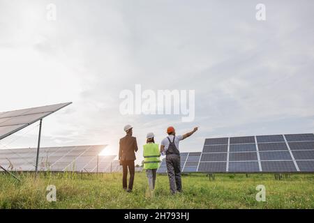 Back view of two inspectors in white helmets walking on solar station with technician in uniform. Multiracial engineers examining work of photovoltaic cells outdoors. Stock Photo