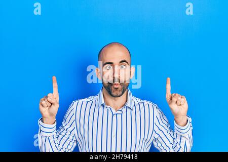 Young bald man pointing up with fingers making fish face with mouth and squinting eyes, crazy and comical. Stock Photo