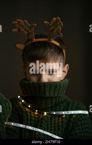 A cute boy in deer horns and a knitted Christmas sweater hides his face